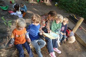 aufmerksame Kinder neben sich sitzend, ließt eine Erzieherin im Garten aus einem Buch über Elektrik vor